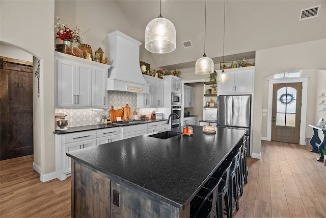 kitchen featuring an island with sink, hanging light fixtures, light wood-type flooring, custom exhaust hood, and white cabinetry