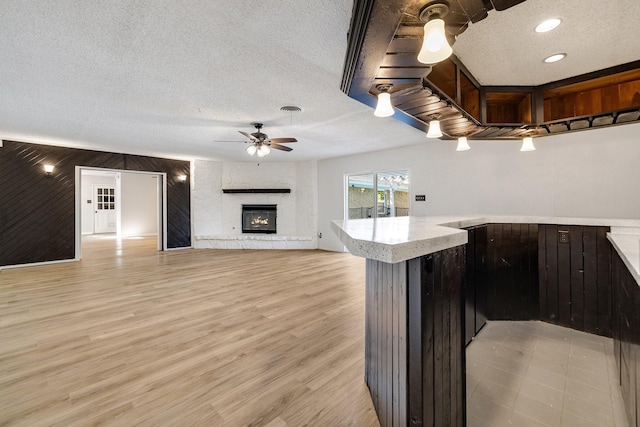 kitchen with ceiling fan, a large fireplace, kitchen peninsula, wood walls, and a textured ceiling