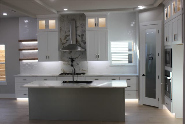 kitchen with wall chimney exhaust hood, white cabinets, light wood-type flooring, and an island with sink