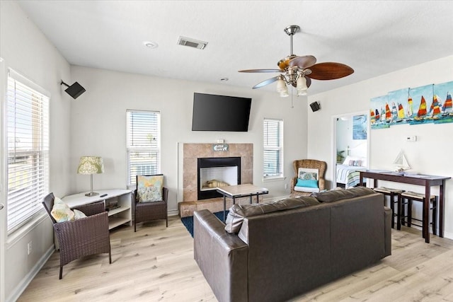 living room with ceiling fan, visible vents, a tiled fireplace, baseboards, and light wood-style floors