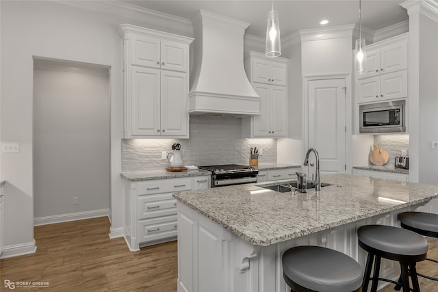 kitchen with appliances with stainless steel finishes, white cabinetry, sink, custom range hood, and a center island with sink