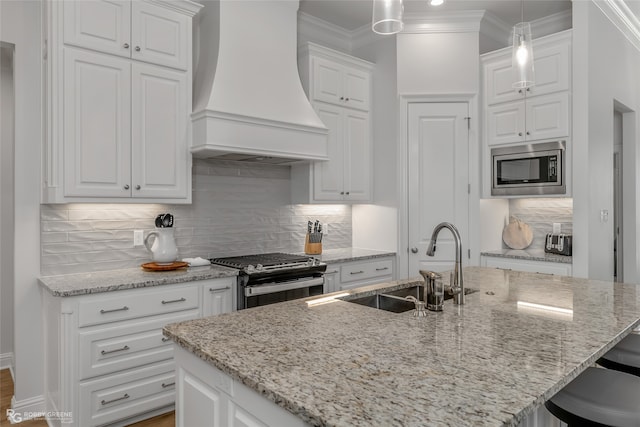 kitchen featuring premium range hood, white cabinetry, hanging light fixtures, appliances with stainless steel finishes, and a kitchen island with sink