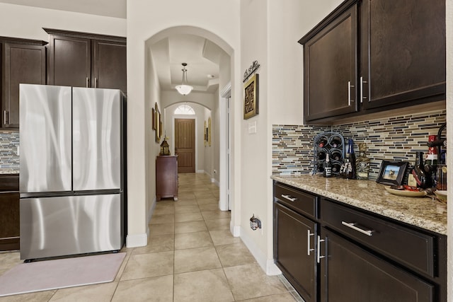 kitchen featuring decorative backsplash, dark brown cabinets, stainless steel refrigerator, light tile patterned flooring, and light stone counters