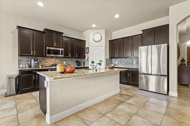 kitchen featuring tasteful backsplash, an island with sink, light stone countertops, dark brown cabinetry, and stainless steel appliances