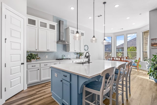 kitchen with sink, white cabinetry, decorative light fixtures, a kitchen island with sink, and wall chimney range hood