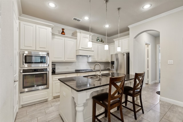 kitchen with stainless steel appliances, dark stone counters, white cabinets, premium range hood, and a center island with sink