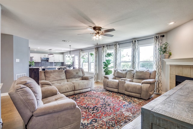 living room featuring a textured ceiling, ceiling fan with notable chandelier, a tiled fireplace, and light wood-type flooring