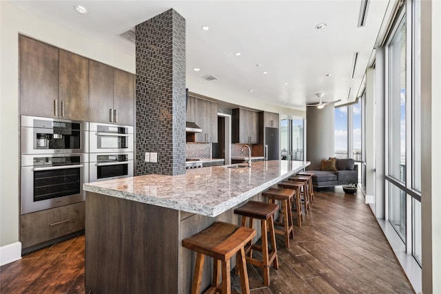 kitchen featuring ventilation hood, sink, a kitchen island with sink, dark brown cabinets, and stainless steel double oven