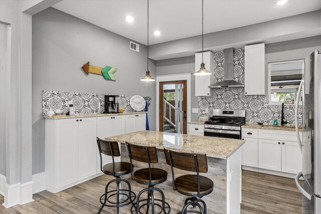 kitchen featuring light stone countertops, appliances with stainless steel finishes, hanging light fixtures, wall chimney exhaust hood, and white cabinets