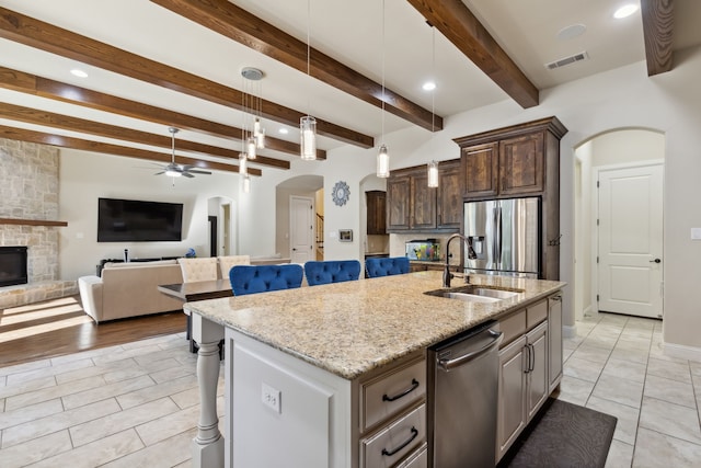 kitchen with a kitchen island with sink, a stone fireplace, sink, beam ceiling, and stainless steel appliances