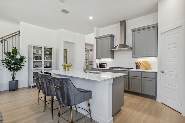 kitchen with a center island with sink, a breakfast bar, light wood-type flooring, wall chimney exhaust hood, and sink