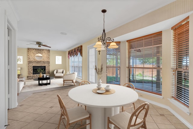 dining area featuring light tile patterned floors, baseboards, crown molding, a brick fireplace, and ceiling fan with notable chandelier