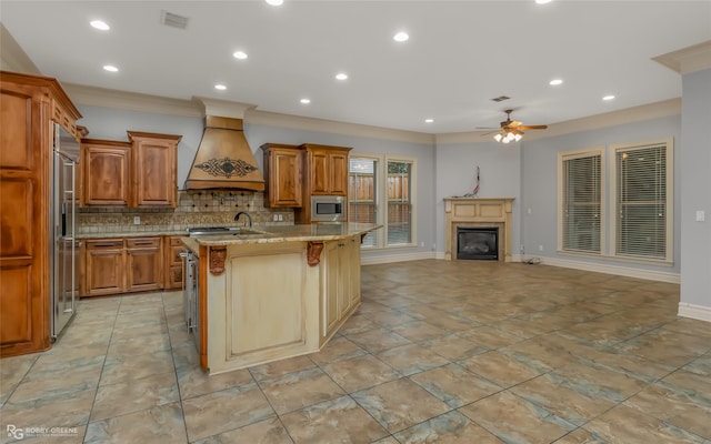 kitchen with a breakfast bar, an island with sink, built in appliances, light stone countertops, and custom range hood