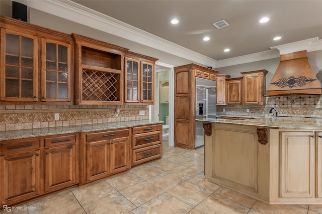 kitchen with built in refrigerator, ornamental molding, light stone countertops, custom exhaust hood, and tasteful backsplash