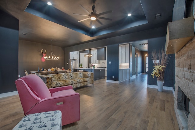 living room featuring a stone fireplace, wood-type flooring, a tray ceiling, and ceiling fan with notable chandelier