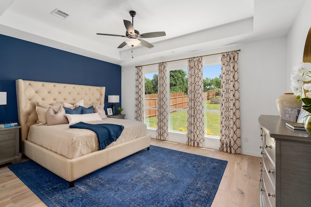 bedroom with a raised ceiling, multiple windows, light wood-type flooring, and ceiling fan