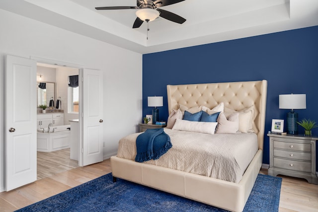 bedroom featuring ensuite bath, a tray ceiling, light wood-type flooring, and ceiling fan