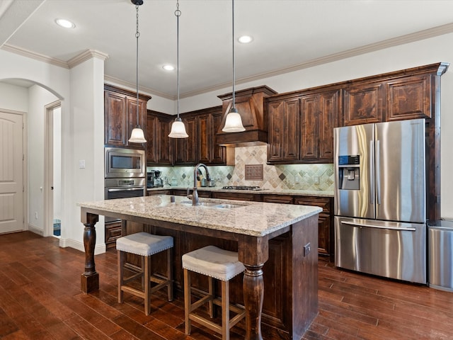 kitchen featuring an island with sink, appliances with stainless steel finishes, light stone countertops, pendant lighting, and premium range hood