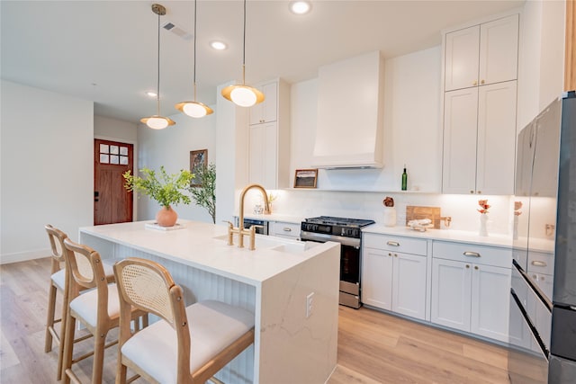 kitchen with an island with sink, hanging light fixtures, stainless steel gas range oven, white cabinets, and light hardwood / wood-style floors