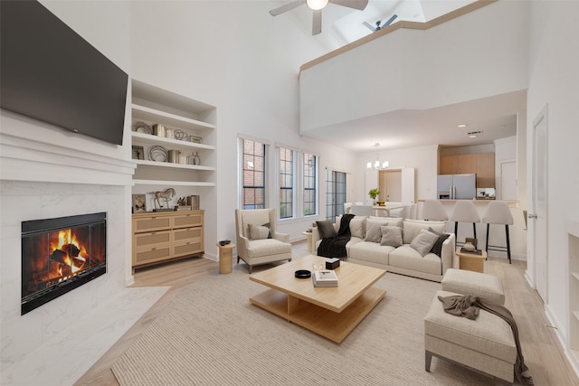 living room featuring built in shelves, ceiling fan with notable chandelier, a tile fireplace, a high ceiling, and light hardwood / wood-style floors