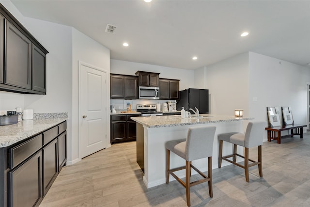 kitchen featuring a center island with sink, appliances with stainless steel finishes, light stone countertops, and light wood-type flooring