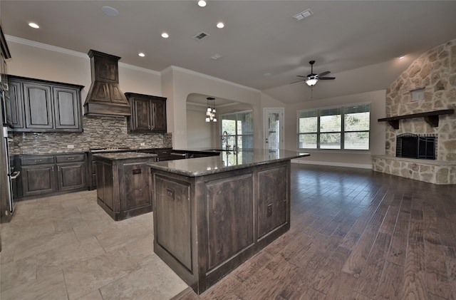 kitchen with tasteful backsplash, a fireplace, custom exhaust hood, light hardwood / wood-style flooring, and a center island with sink