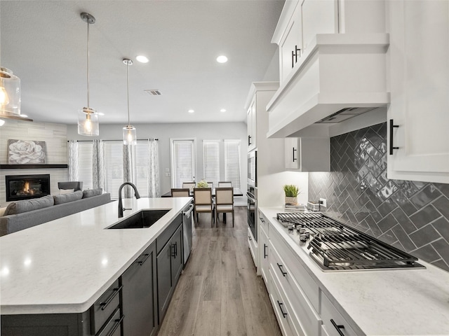 kitchen with stainless steel appliances, custom exhaust hood, a glass covered fireplace, white cabinetry, and a sink