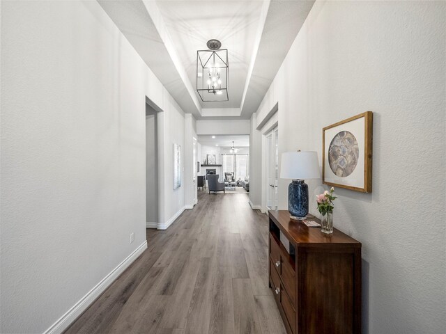 hall featuring baseboards, wood finished floors, a tray ceiling, a textured wall, and a notable chandelier