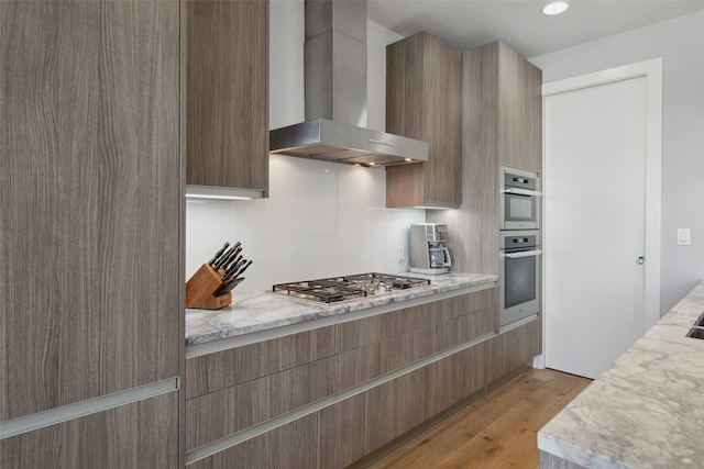 kitchen with light stone counters, wall chimney range hood, stainless steel appliances, and light wood-type flooring