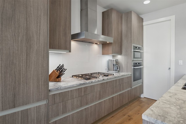 kitchen featuring light stone countertops, appliances with stainless steel finishes, light wood finished floors, wall chimney exhaust hood, and modern cabinets