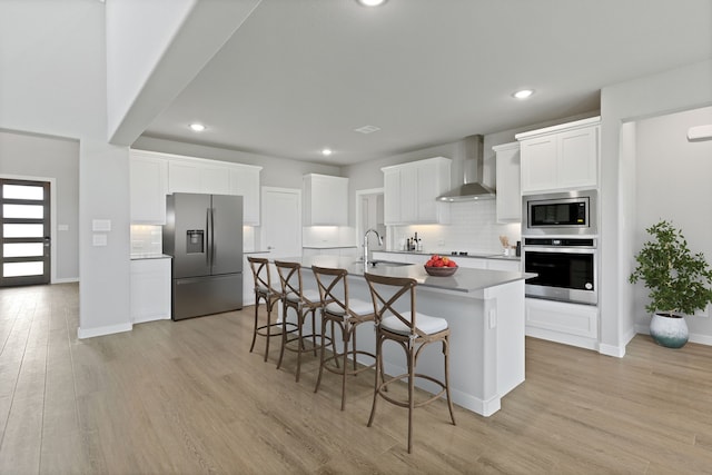 kitchen featuring wall chimney range hood, light hardwood / wood-style flooring, a center island with sink, white cabinets, and appliances with stainless steel finishes