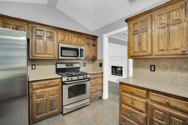 kitchen featuring lofted ceiling, backsplash, appliances with stainless steel finishes, a fireplace, and light stone counters