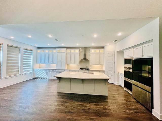 kitchen with wall chimney range hood, a center island with sink, backsplash, white cabinetry, and dark hardwood / wood-style floors