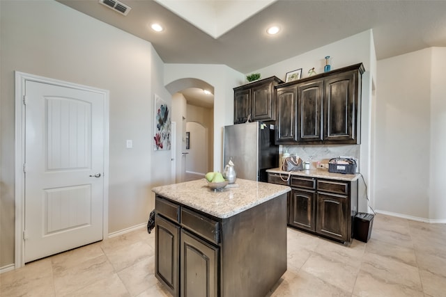 kitchen with tasteful backsplash, a kitchen island, dark brown cabinets, stainless steel fridge, and light stone counters