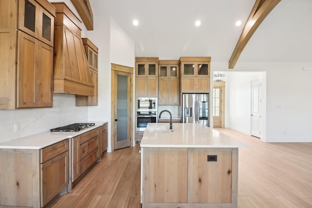 kitchen with light wood-type flooring, custom range hood, stainless steel appliances, a kitchen island with sink, and beam ceiling