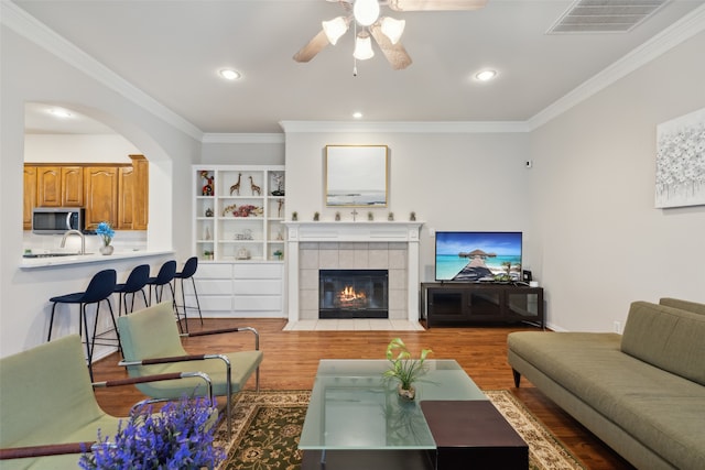 living room featuring a tiled fireplace, ceiling fan, wood-type flooring, ornamental molding, and sink