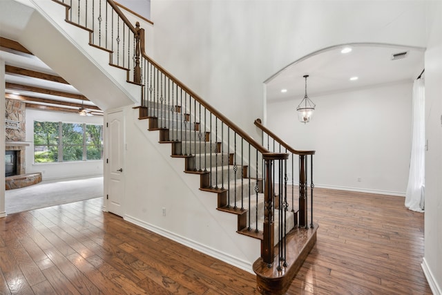 staircase with a stone fireplace, an inviting chandelier, beam ceiling, and wood-type flooring