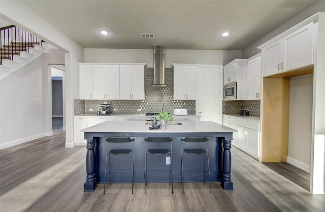 kitchen featuring stainless steel microwave, wall chimney exhaust hood, white cabinets, and a kitchen island with sink