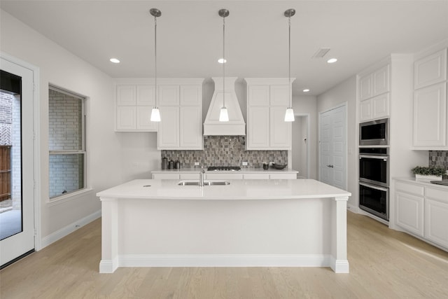 kitchen featuring sink, white cabinetry, custom range hood, stainless steel appliances, and a kitchen island with sink