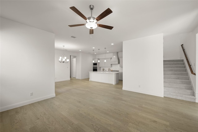 unfurnished living room featuring recessed lighting, ceiling fan with notable chandelier, baseboards, stairway, and light wood finished floors