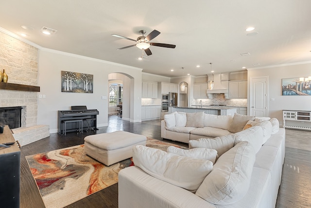 living room featuring ceiling fan, dark wood-type flooring, crown molding, and a stone fireplace