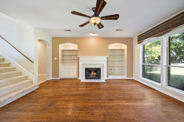 unfurnished living room with ceiling fan, built in features, wood-type flooring, and a tile fireplace