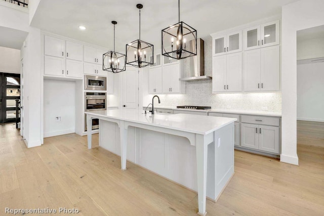 kitchen with wall chimney range hood, stainless steel microwave, a center island with sink, pendant lighting, and white cabinetry