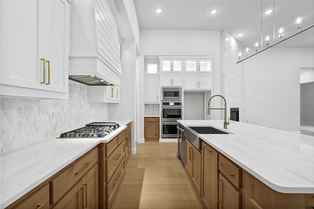 kitchen with premium range hood, white cabinets, sink, stainless steel appliances, and a center island with sink