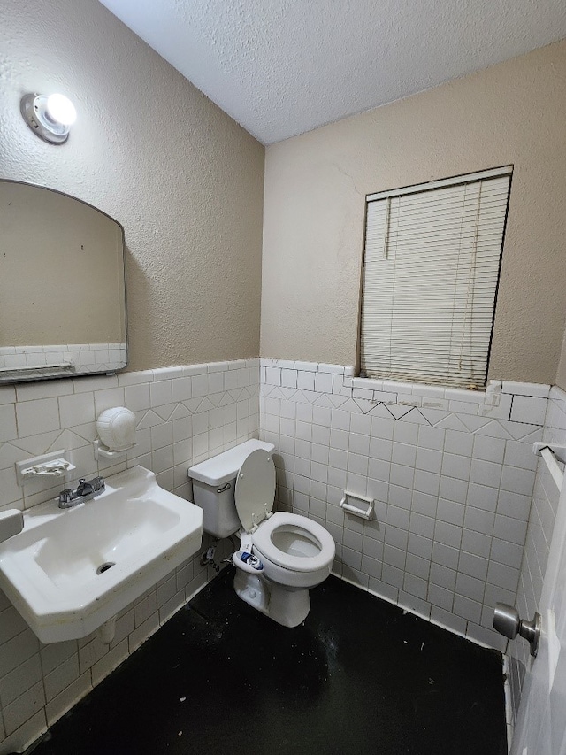 half bathroom with toilet, tile walls, a textured ceiling, a wainscoted wall, and a sink