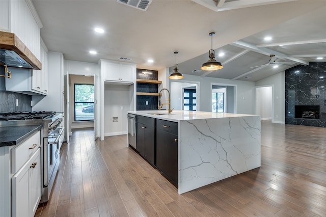 kitchen featuring white cabinetry, appliances with stainless steel finishes, an island with sink, hanging light fixtures, and premium range hood