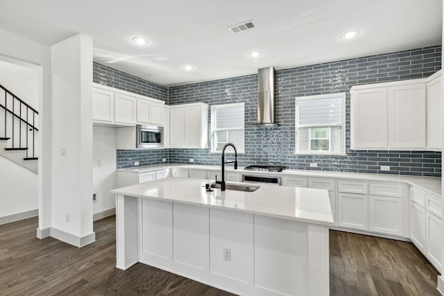 kitchen with white cabinets, appliances with stainless steel finishes, a kitchen island with sink, dark hardwood / wood-style floors, and wall chimney exhaust hood