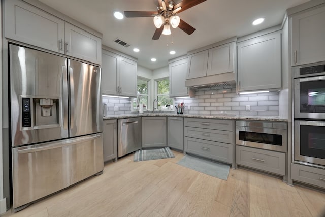 kitchen with appliances with stainless steel finishes, light wood-type flooring, backsplash, gray cabinets, and light stone counters