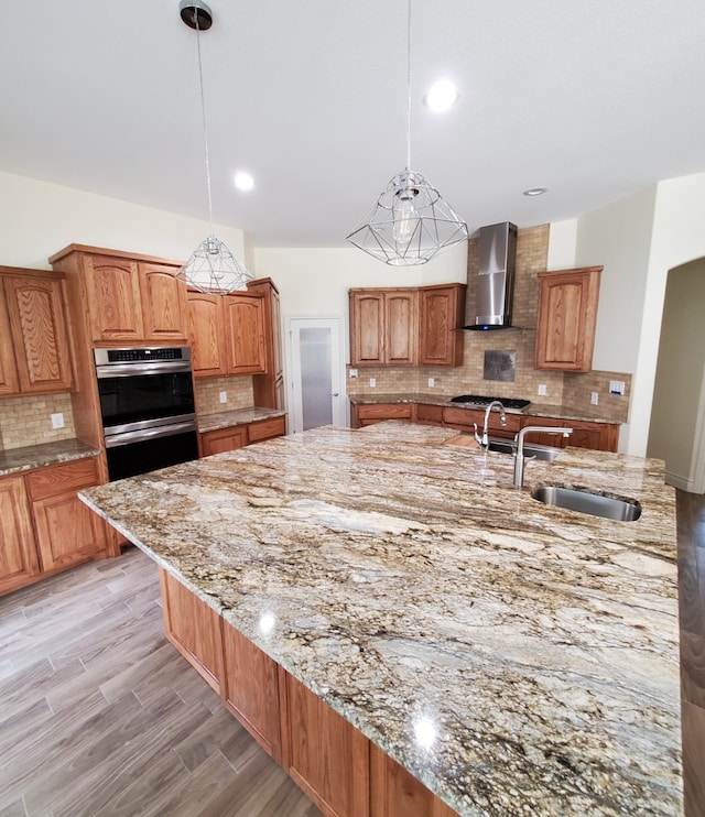 kitchen with wall chimney range hood, backsplash, light stone countertops, pendant lighting, and stainless steel appliances