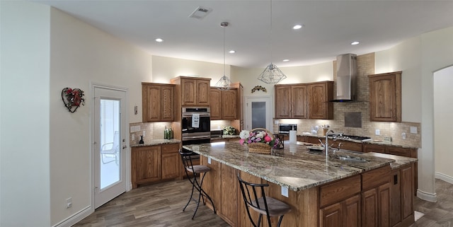 kitchen with a sink, visible vents, wall chimney range hood, brown cabinets, and a center island with sink